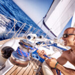 Muž na palube plachetnice, usmievajúci sa a manipulujúci s lanami, symbolizujúci skúsenosti kapitána na mori.