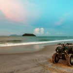 Štvorkolka zaparkovaná na piesočnatej pláži pri mori s výhľadom na ostrov pri západe slnka.