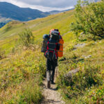 Turista s veľkým a ťažkým batohom na chrbte v pevnej členkovej turistickej obuvi prechádzajúci náročným terénom.